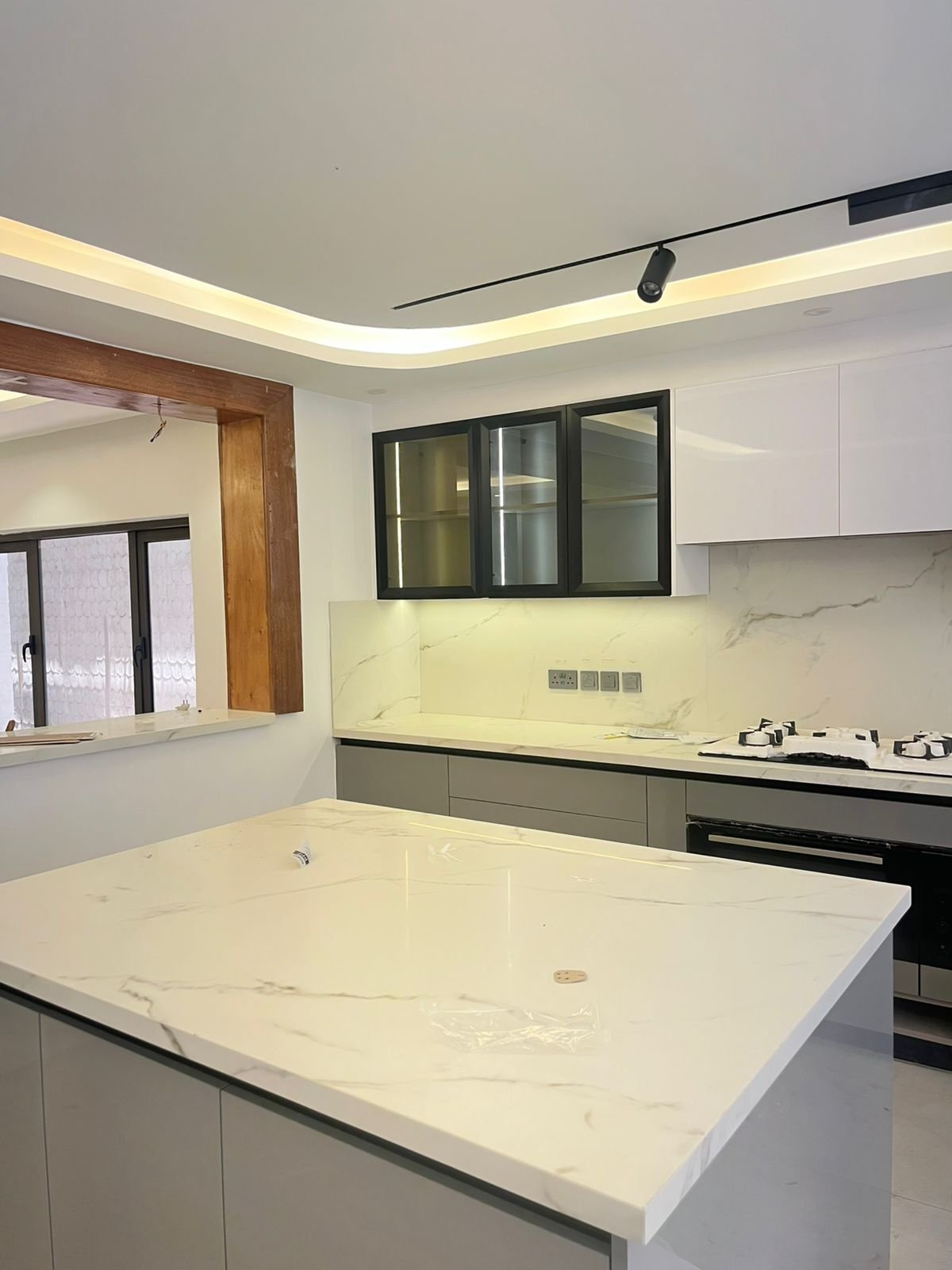 Modern kitchen with white marble island counter, black lower cabinets, glass-front upper cabinets, and cream-colored cove ceiling lighting