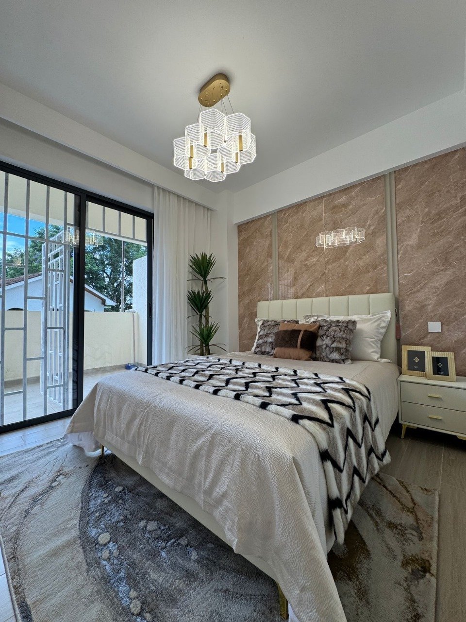 Modern bedroom with queen bed, geometric patterned bedding, textured brown accent wall, pendant light fixture, and glass door opening to balcony