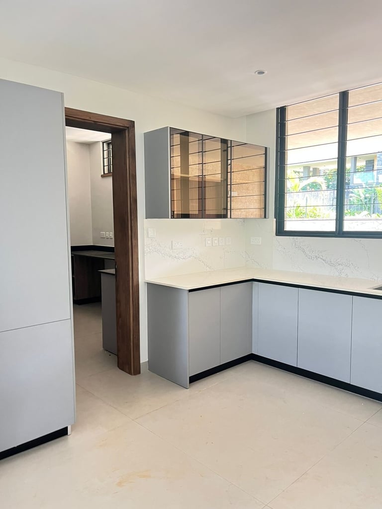 Modern kitchen interior with white cabinetry, glass-fronted upper cabinets, beige countertops, large window with natural light, and open doorway to adjacent room