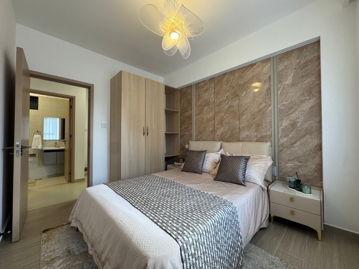 Modern bedroom with queen bed, padded headboard, wooden wardrobe, nightstands, flower-shaped ceiling lamp, and ensuite bathroom visible through open door