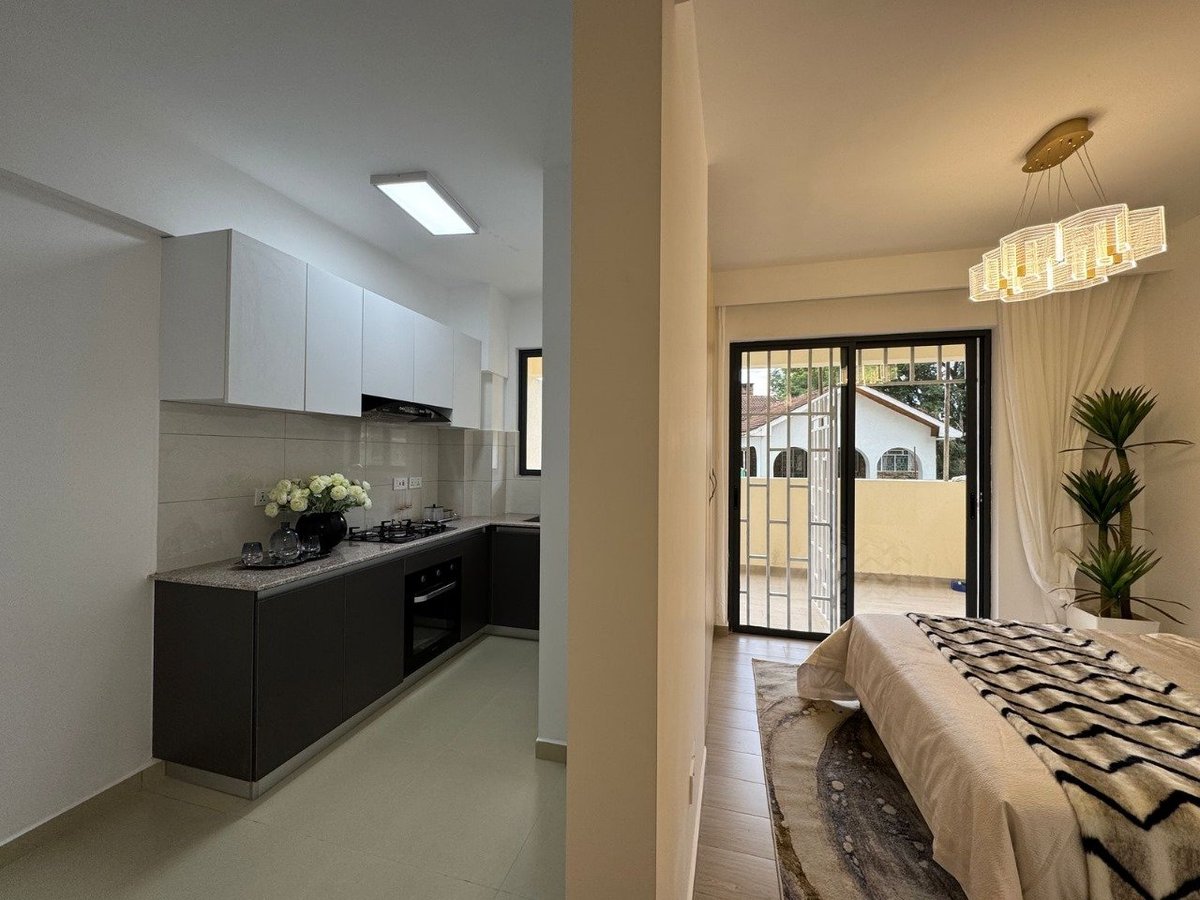 Modern apartment interior with open floor plan showing kitchen with white upper cabinets and black lower cabinets on left, bedroom area on right with glass doors leading to balcony