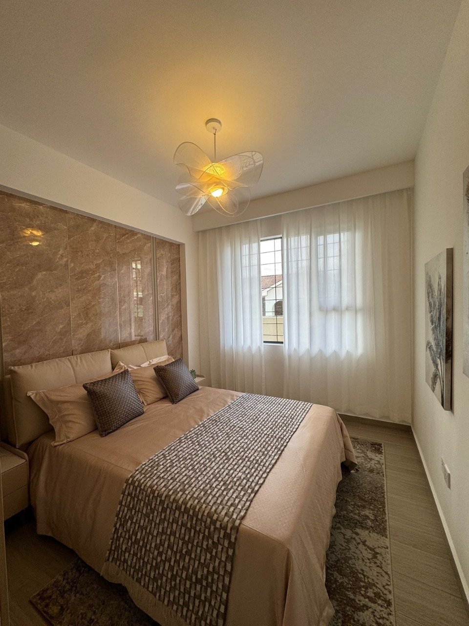 Modern bedroom with beige bed, textured headboard, glowing flower-shaped ceiling light, and sheer curtains over a window